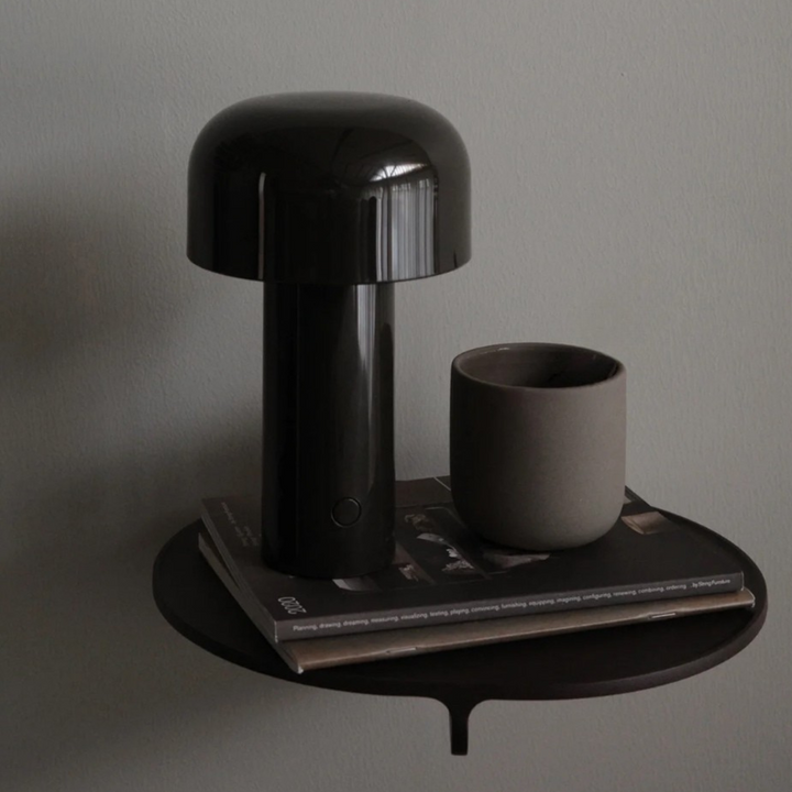 A black cordless mushroom lamp on a floating wall-mounted shelf, styled with a neutral-toned cup and magazine in a bedroom.