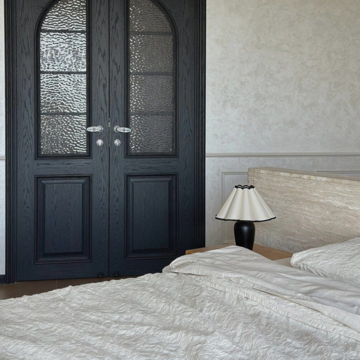 Black table lamp with a unique scalloped shade on a bedside table in a minimalist bedroom with French-inspired decor.