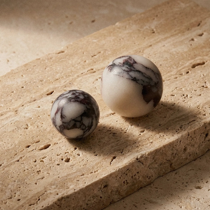 Two Calacatta Viola marble knobs resting on a travertine block, warm afternoon light showing the contrast between the stones.