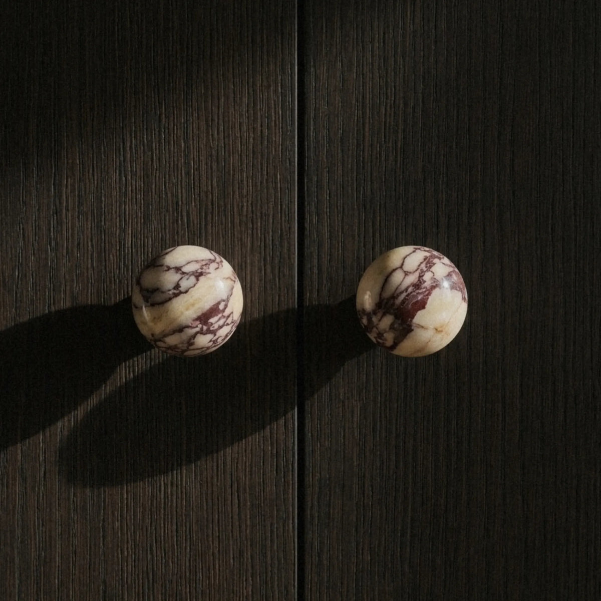 Two Calacatta Viola marble knobs on a dark wood cabinet cream base with deep violet veining, strong side light.