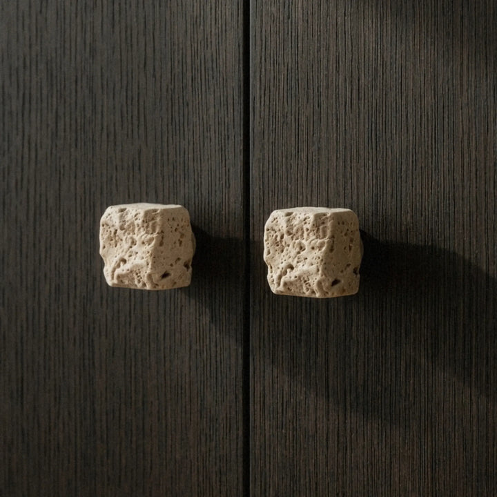 Close-up of two chunky travertine cupboard knobs on dark wood uneven edges and porous texture showing the raw stone character.