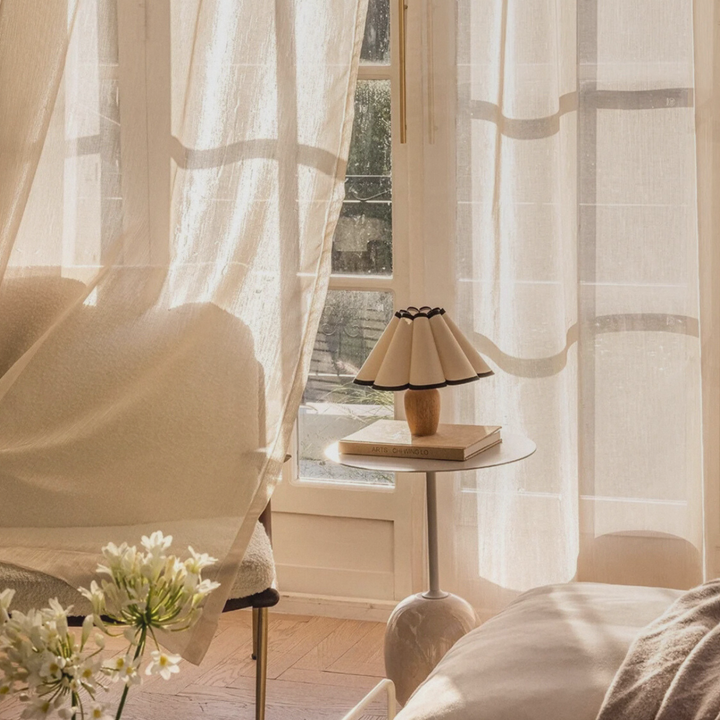 A French-inspired table lamp with a pleated shade and bold trim, styled next to a cream armchair with sheer curtains and natural light.