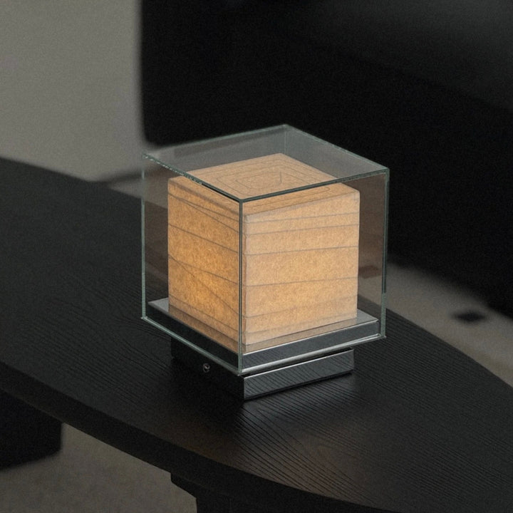 glass table lamp with a cube form placed on a dark surface emitting soft light.