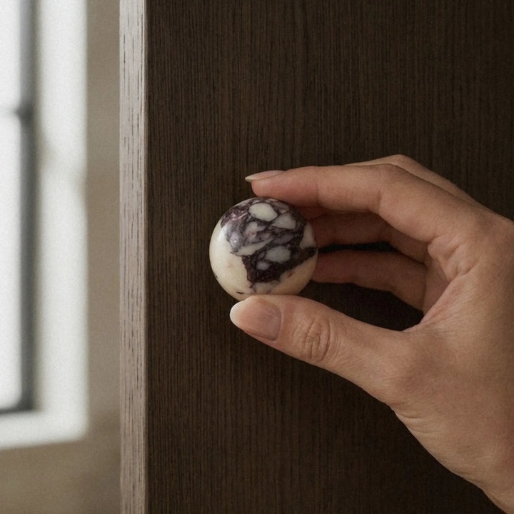 Hand picking up a Calacatta Viola marble knob near a bright window, violet veining clearly visible against the cream surface.