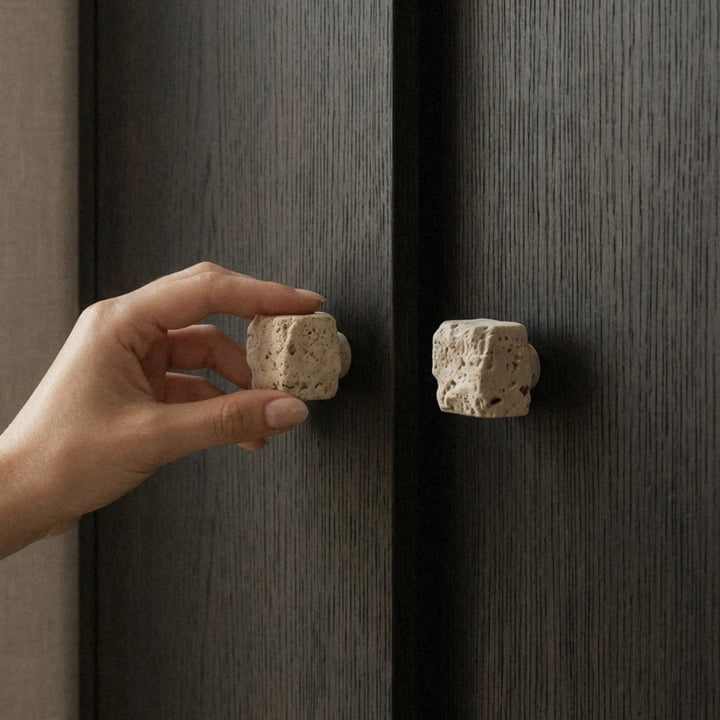 Hand closing a dark cabinet door using a rough travertine cube knob, warm light catching the stone's surface.