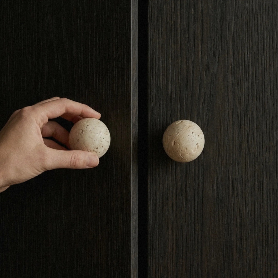 Hand pulling open a cabinet door by a round travertine knob, soft natural light from the side.