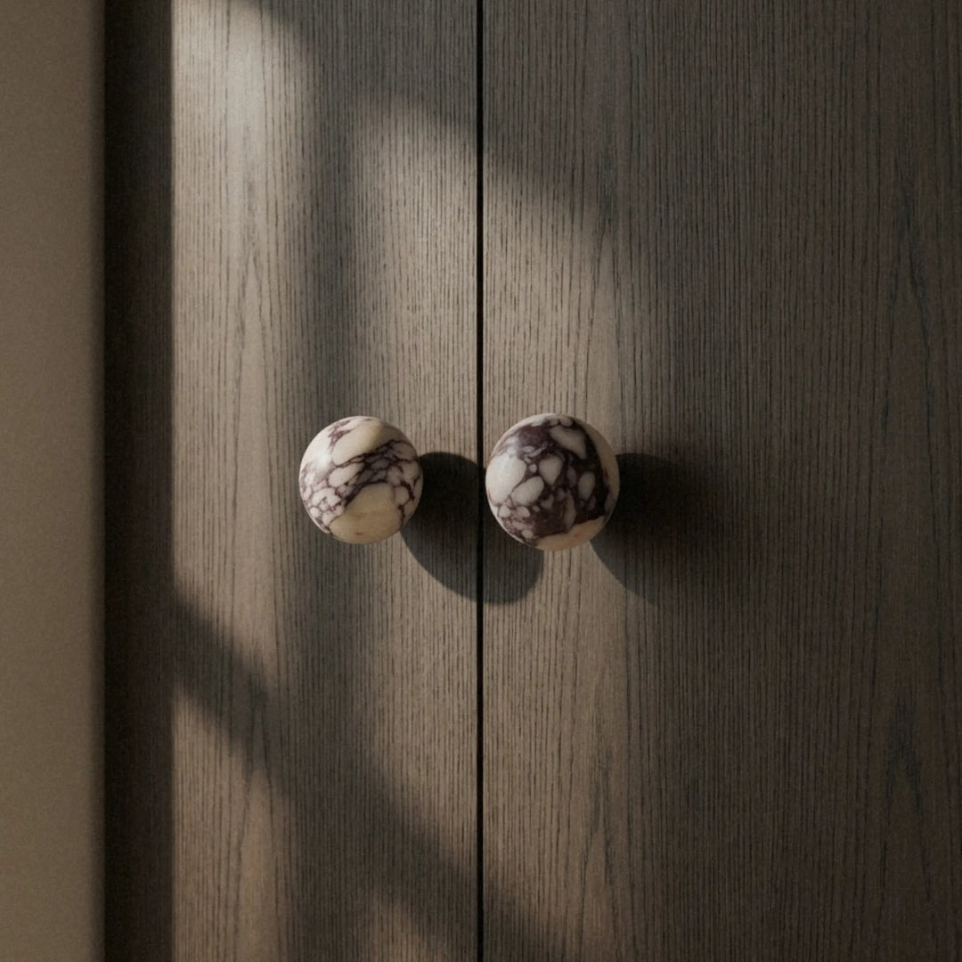 Calacatta Viola cabinet knob seen through a narrow gap between two dark doors, soft light from behind.