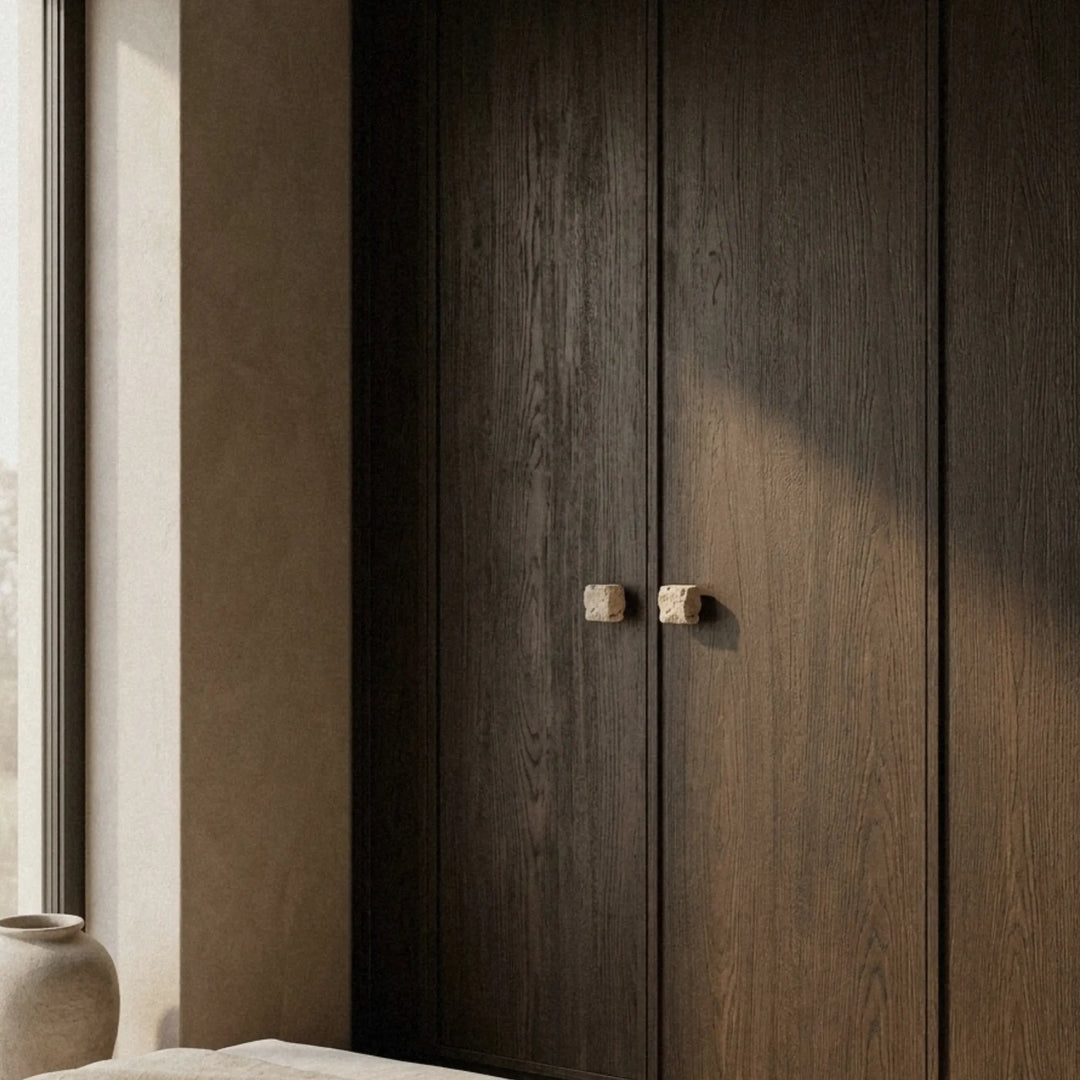 Travertine cube knob on a tall dark wardrobe door, seen from a distance in a quiet room with soft window light.