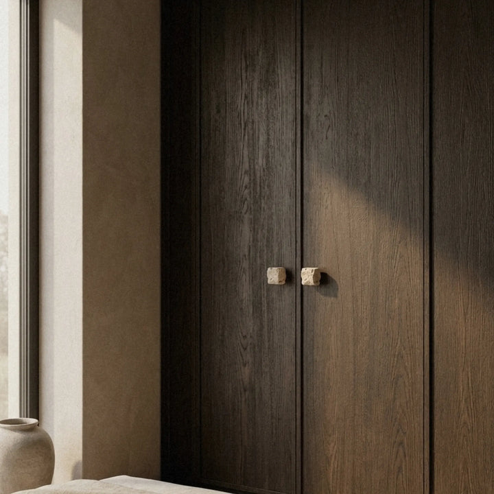 Travertine cube knob on a tall dark wardrobe door, seen from a distance in a quiet room with soft window light.