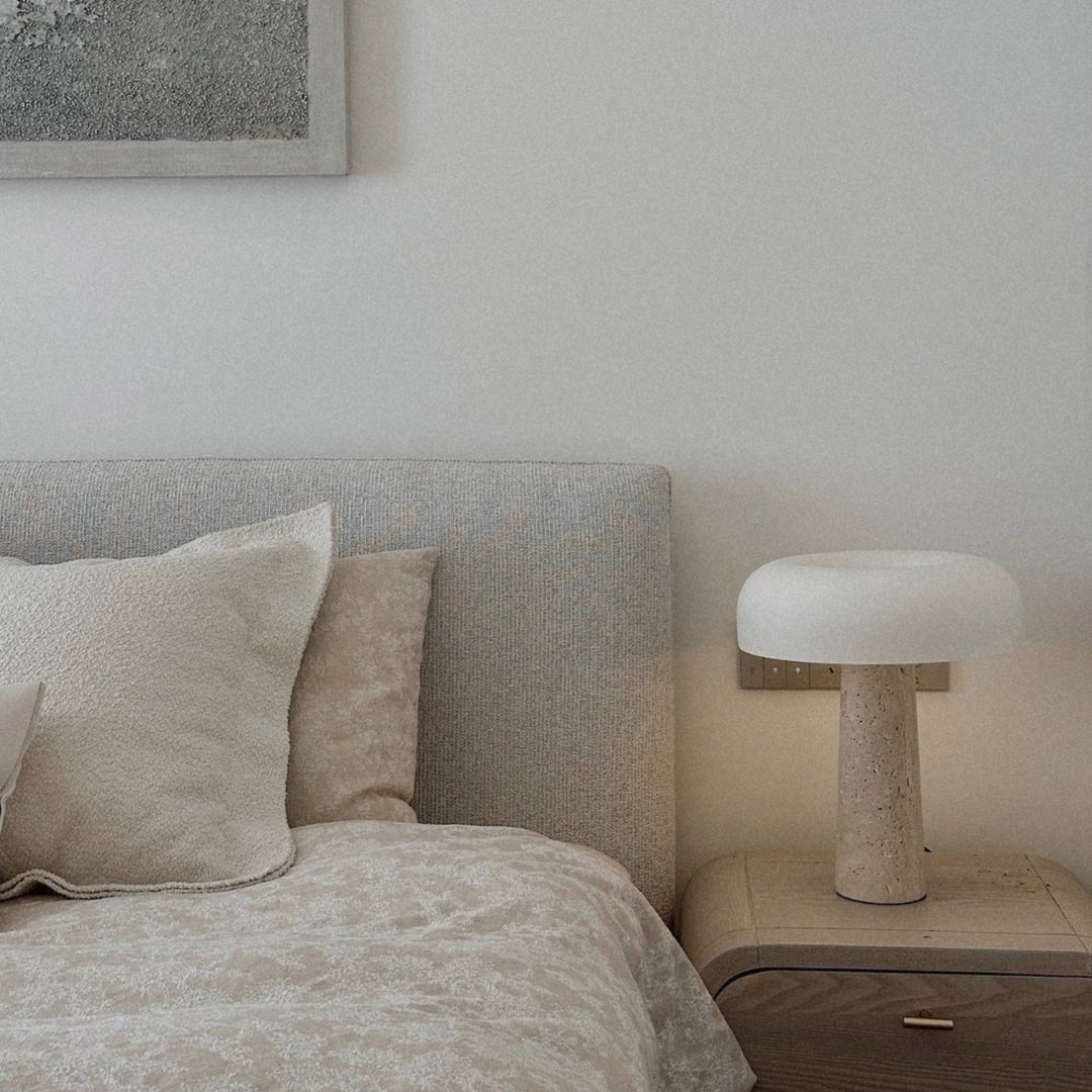 minimalist stone table lamp placed on a bedside table beside a soft neutral bed setting.