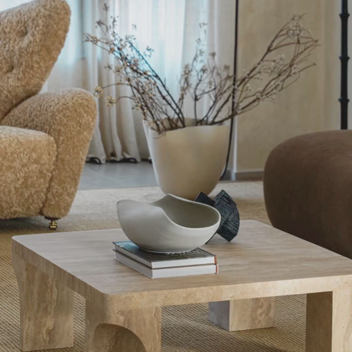 Modern ceramic bowl with bold organic curves placed on a minimalist travertine coffee table in a cozy neutral-toned living room.