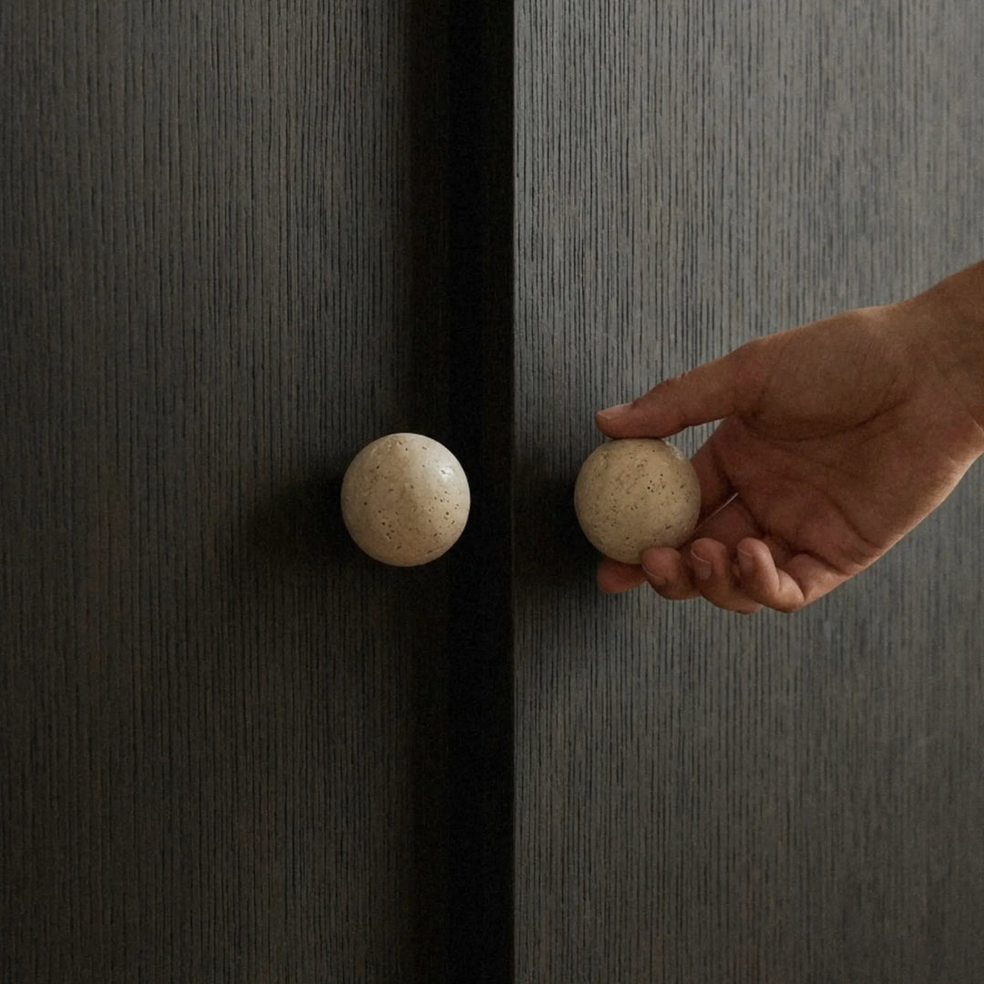 A hand gently reaching toward a travertine knob on a dark cabinet door in a dimly lit room.