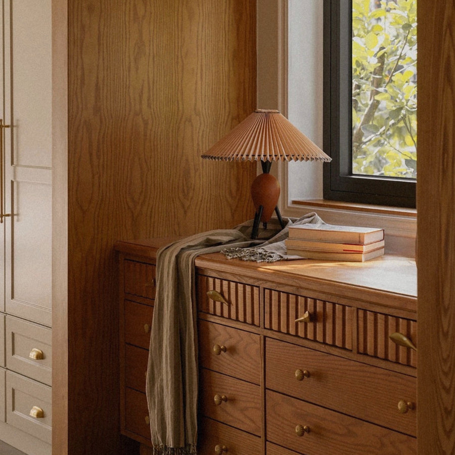 pleated table lamp with wooden base placed on a dresser beside a window in a warm bedroom interior.