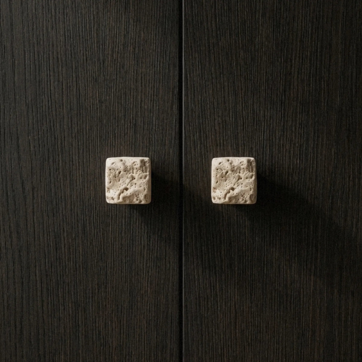 Two rough-cut travertine cube knobs on a dark oak cabinet, natural pitting visible across the stone surface.