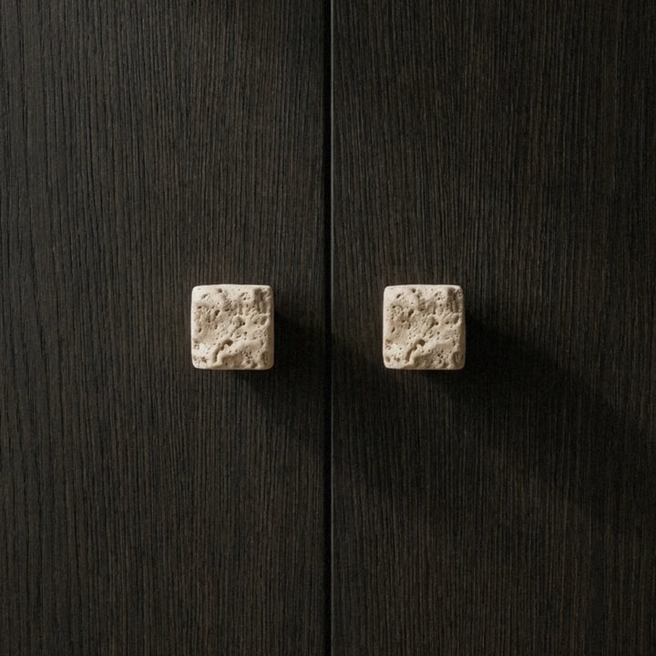 Two rough-cut travertine cube knobs on a dark oak cabinet, natural pitting visible across the stone surface.