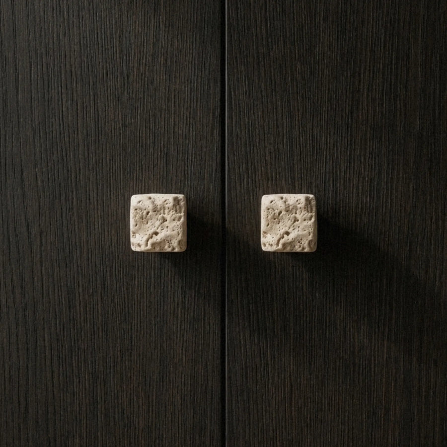 Two rough-cut travertine cube knobs on a dark oak cabinet, natural pitting visible across the stone surface.