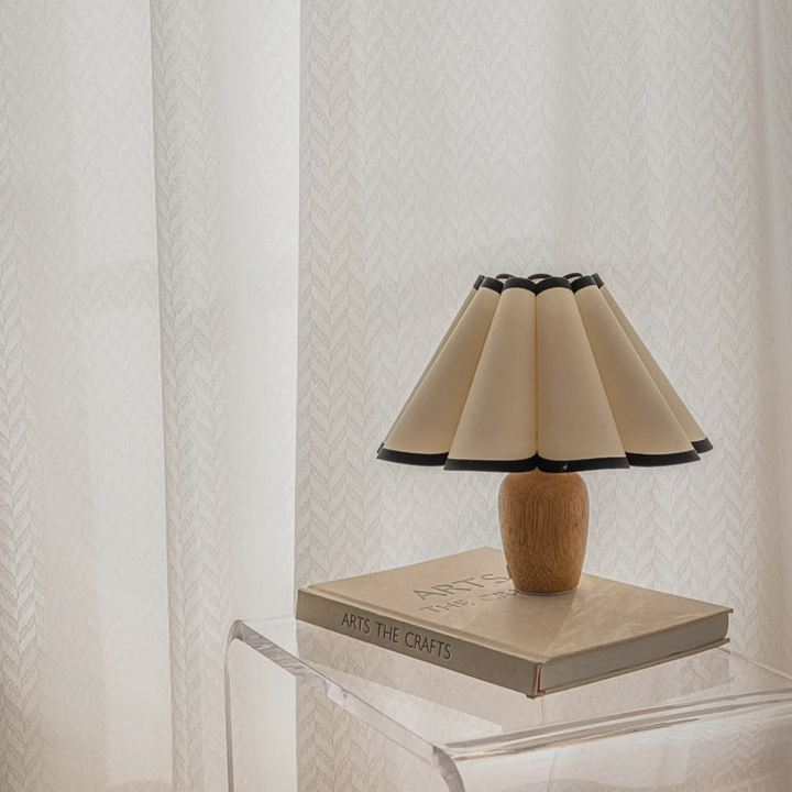 Small table lamp with a pleated shade and natural wood base, placed on a clear acrylic table with a minimal and modern vibe.