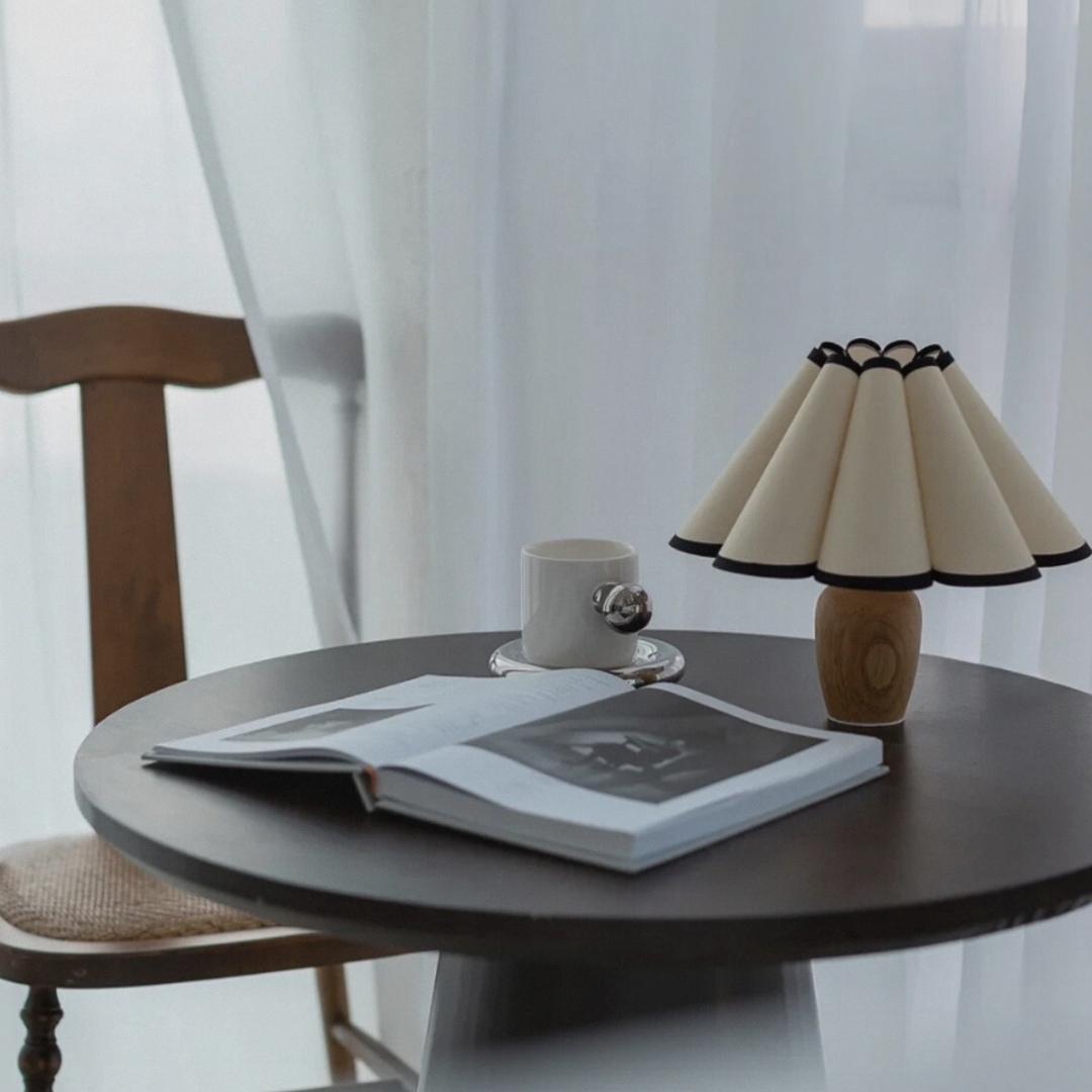 Modern table lamp with a sculptural pleated shade and black trim, placed on a dark round table by a wooden chair in a Scandinavian-inspired setting.