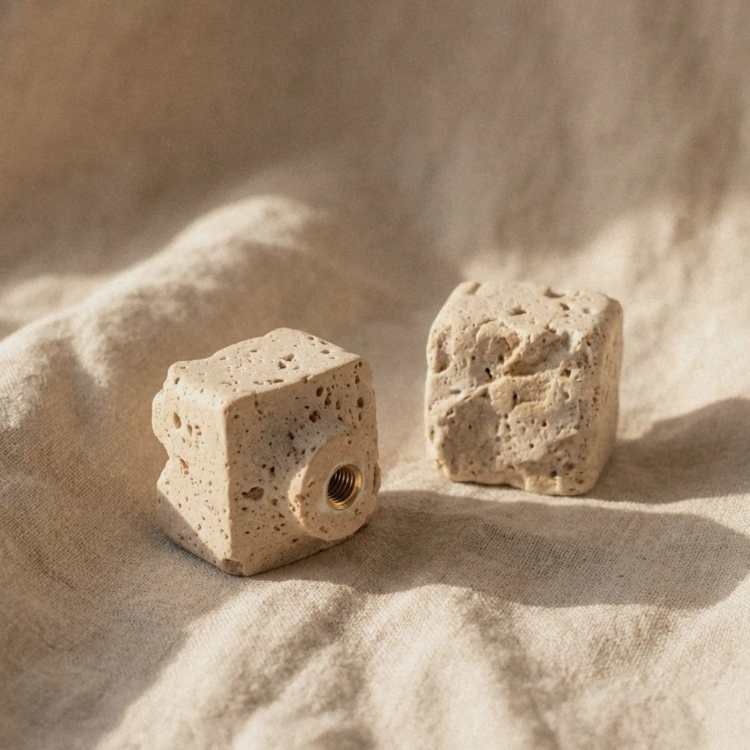 Two raw travertine cube knobs on linen cloth, afternoon shadow cutting across the porous stone faces.