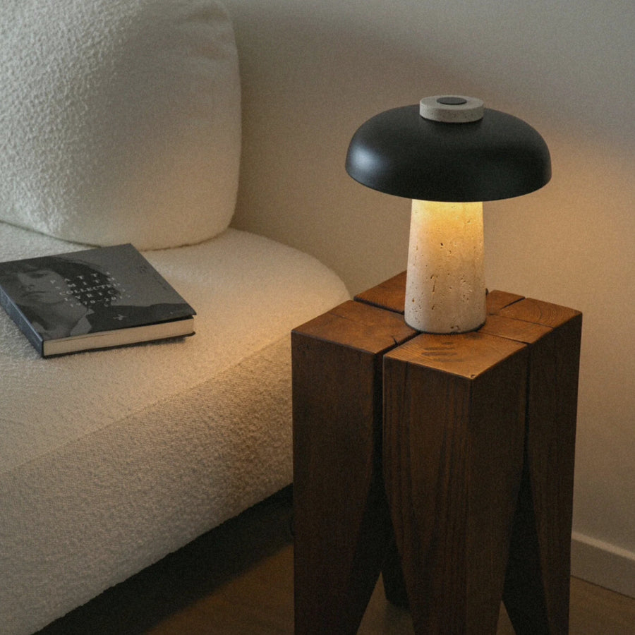 travertine stone table lamp with a dark iron shade resting on a wooden side table beside a sofa.